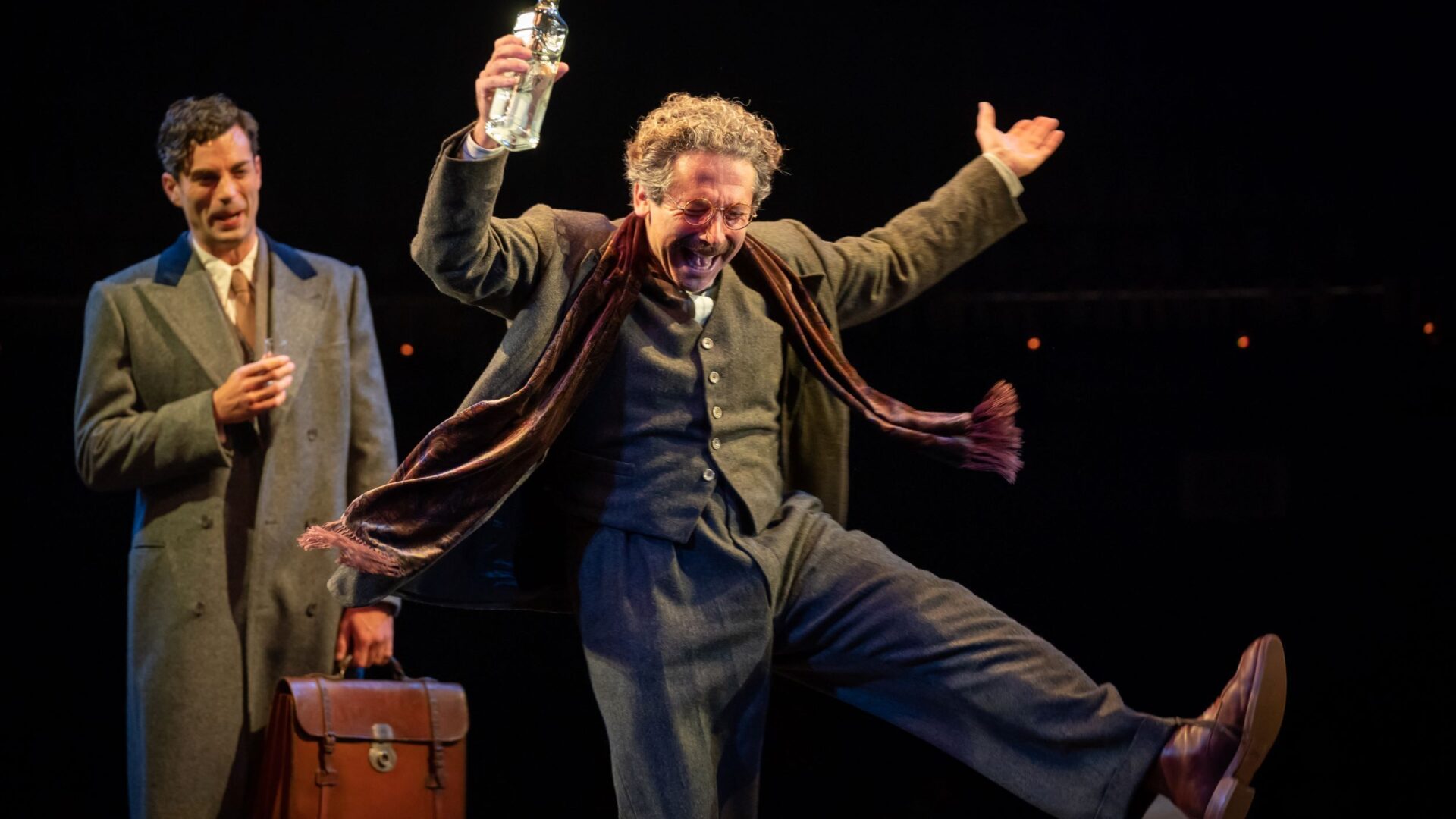 Production photo from cabaret. Actor in a suit in the background holding a drink and smiling; another in the foreground in a suit with a scarf dancing, celebrating.