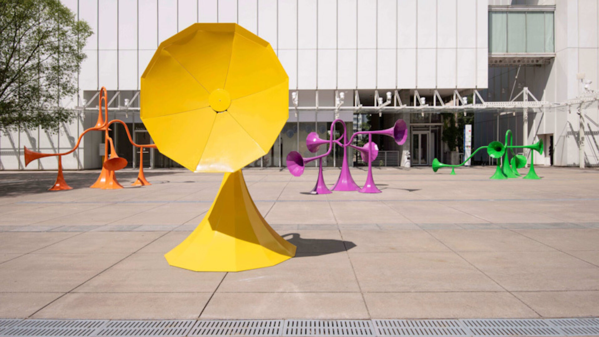Big yellow horn in an open space with orange, purple and green interactive sound horns as well as a white building in the background - Yuri Suzuki Sonic Playground