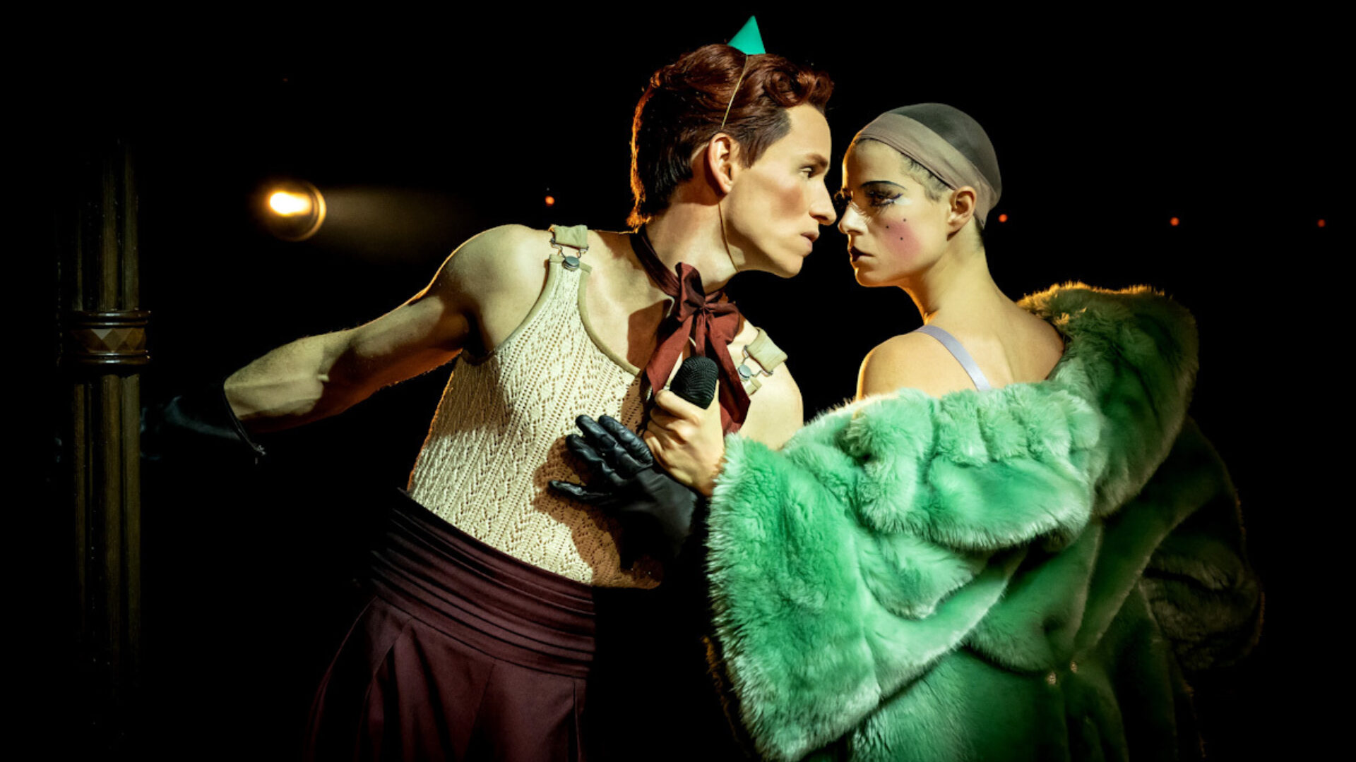 Production photo from the 2021 London production of Cabaret. On the left, the Emcee wearing a beige tank top and brown pants; Sally on the left in a green fur coat.
