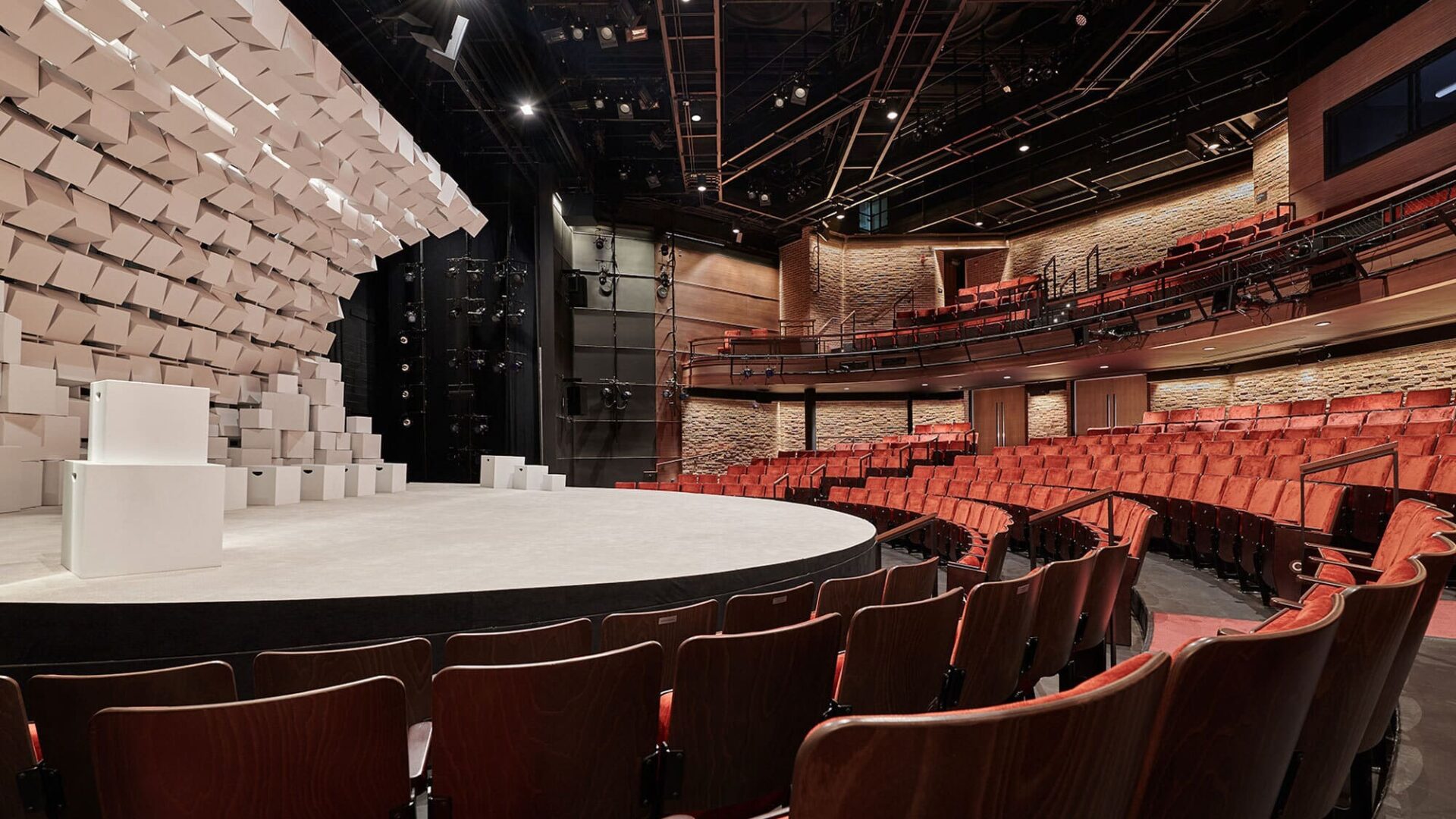 The Roundhouse Theatre stage to the left with a curved wall with white cubes, to the right the wooden seats and the exposed brick wall in the back and the catwalk above.