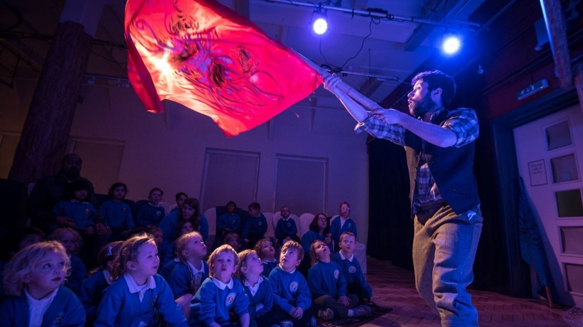 A dynamic storytelling session in a dimly lit room with a performer waving a red flag with intricate designs, captivating an audience of young children in school uniforms, who look on with expressions of wonder and engagement.