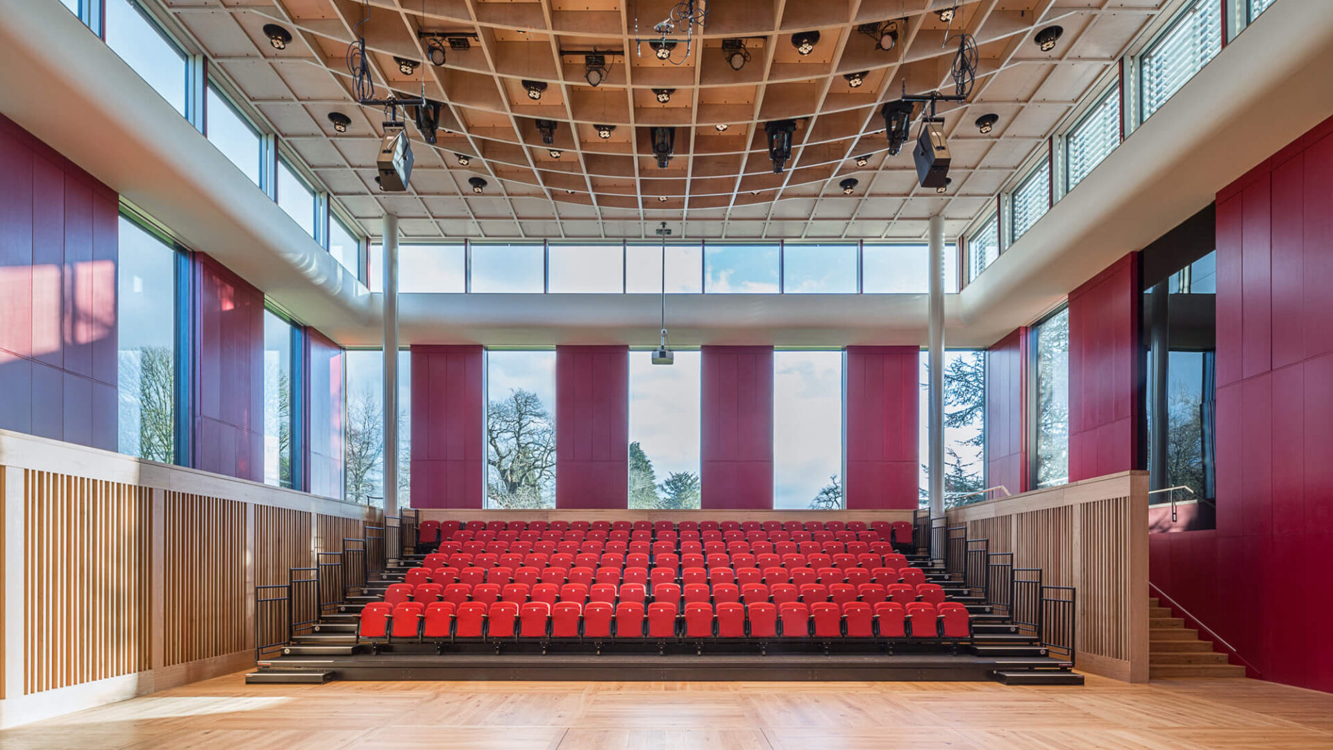 Eavis Hall view from the stage with empty red seats and large dark red wall panels and windows as well as the exposed wooden beams in the roof with lighting equipment