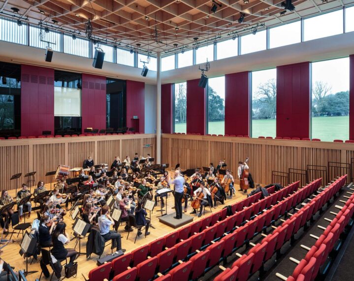 Side view of students playing musical instruments rehearsing in Eavis Hall, with wooden panels, dark red walls and large windows with exposed structural beams in the roof